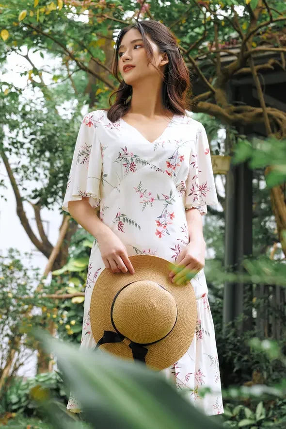 Woman in a white floral modern cheongsam dress holding a straw hat, standing in a garden.