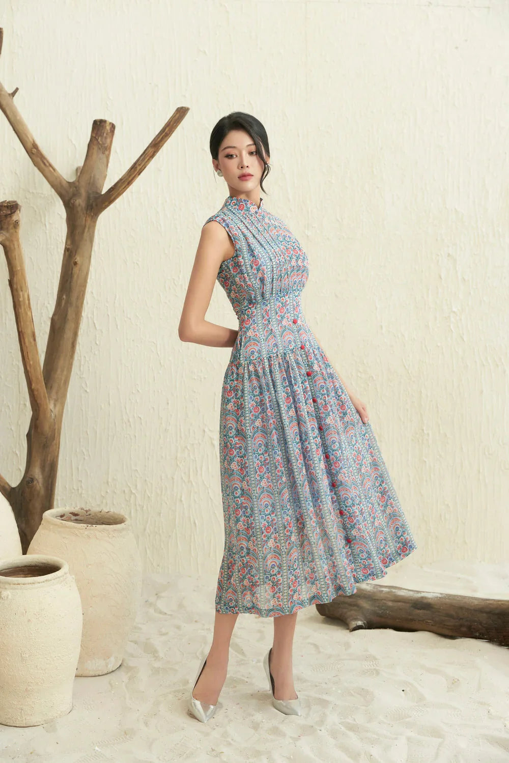 Woman wearing a sleeveless modern cheongsam dress with blue and pink floral pattern, standing indoors on sand by pottery and driftwood.