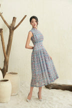 Woman wearing a sleeveless modern cheongsam dress with blue and pink floral pattern, standing indoors on sand by pottery and driftwood.