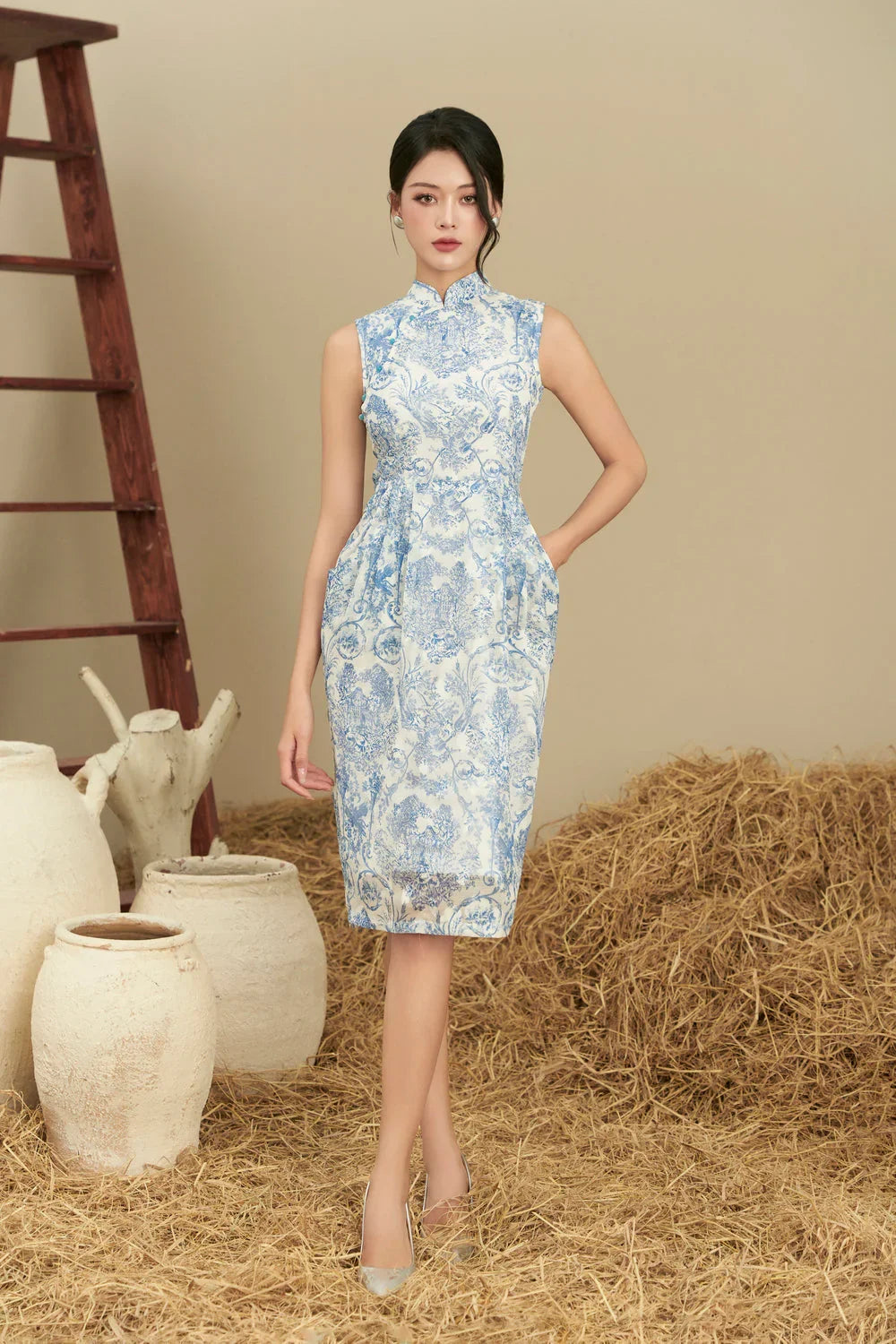 Woman in sleeveless blue and white cheongsam dress with floral pattern, standing on straw