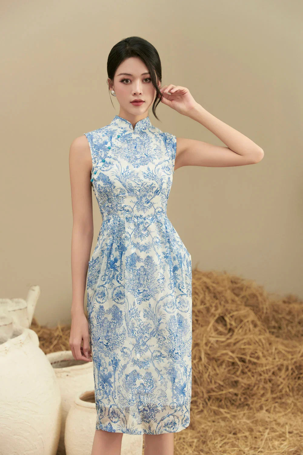 Woman wearing a sleeveless blue and white floral cheongsam dress, standing indoors.