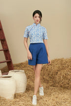 Woman in modern blue floral cheongsam top and blue shorts, standing on straw indoors