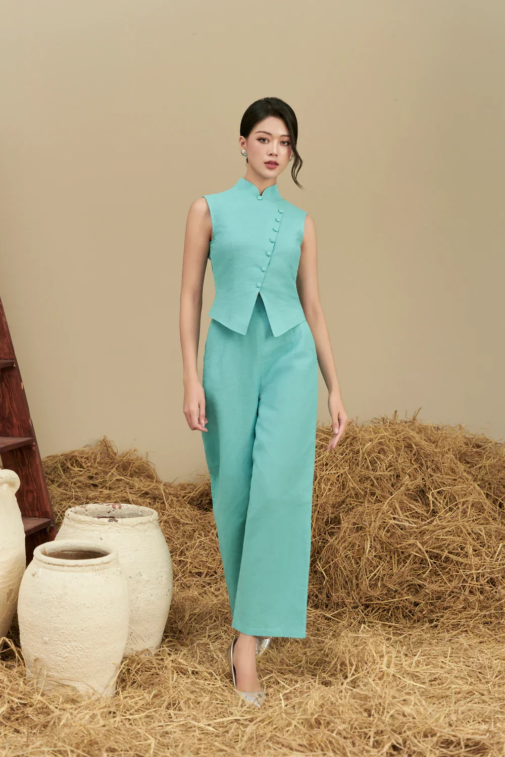 Woman wearing sleeveless teal modern cheongsam jumpsuit with wide leg pants, standing indoors