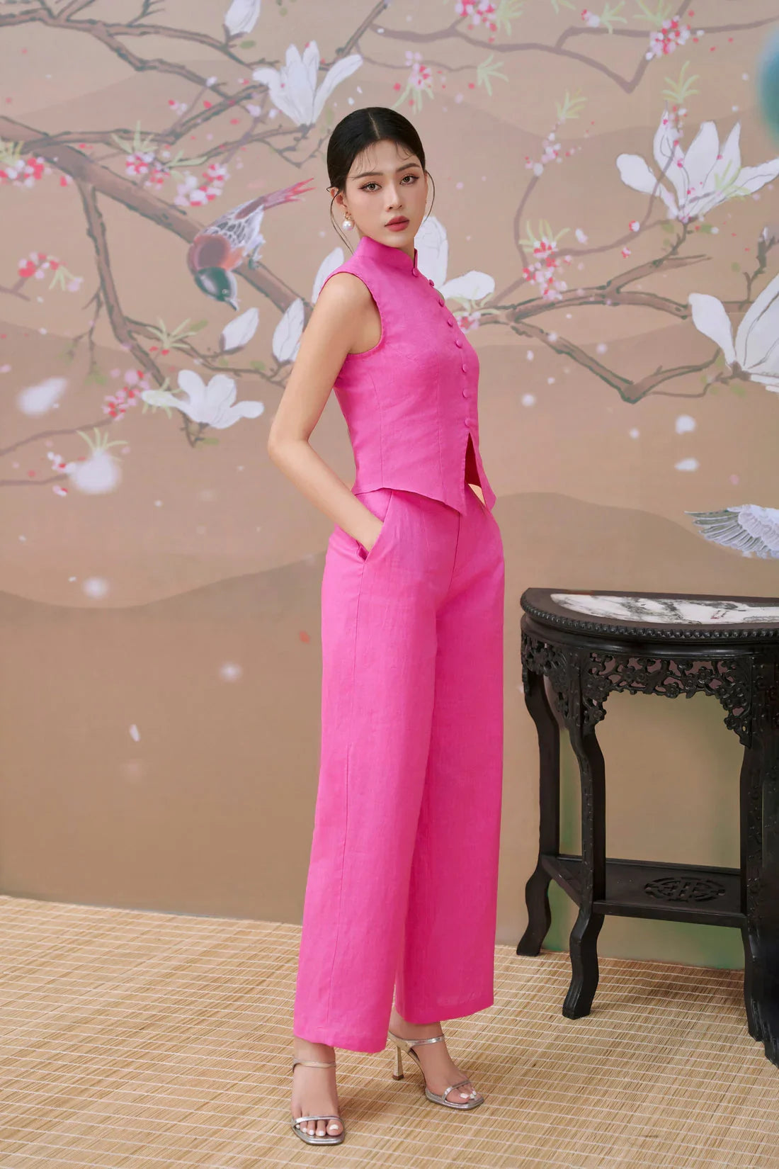 Woman in a modern pink cheongsam-inspired set with sleeveless top and wide-leg pants, standing in front of a floral Asian backdrop.