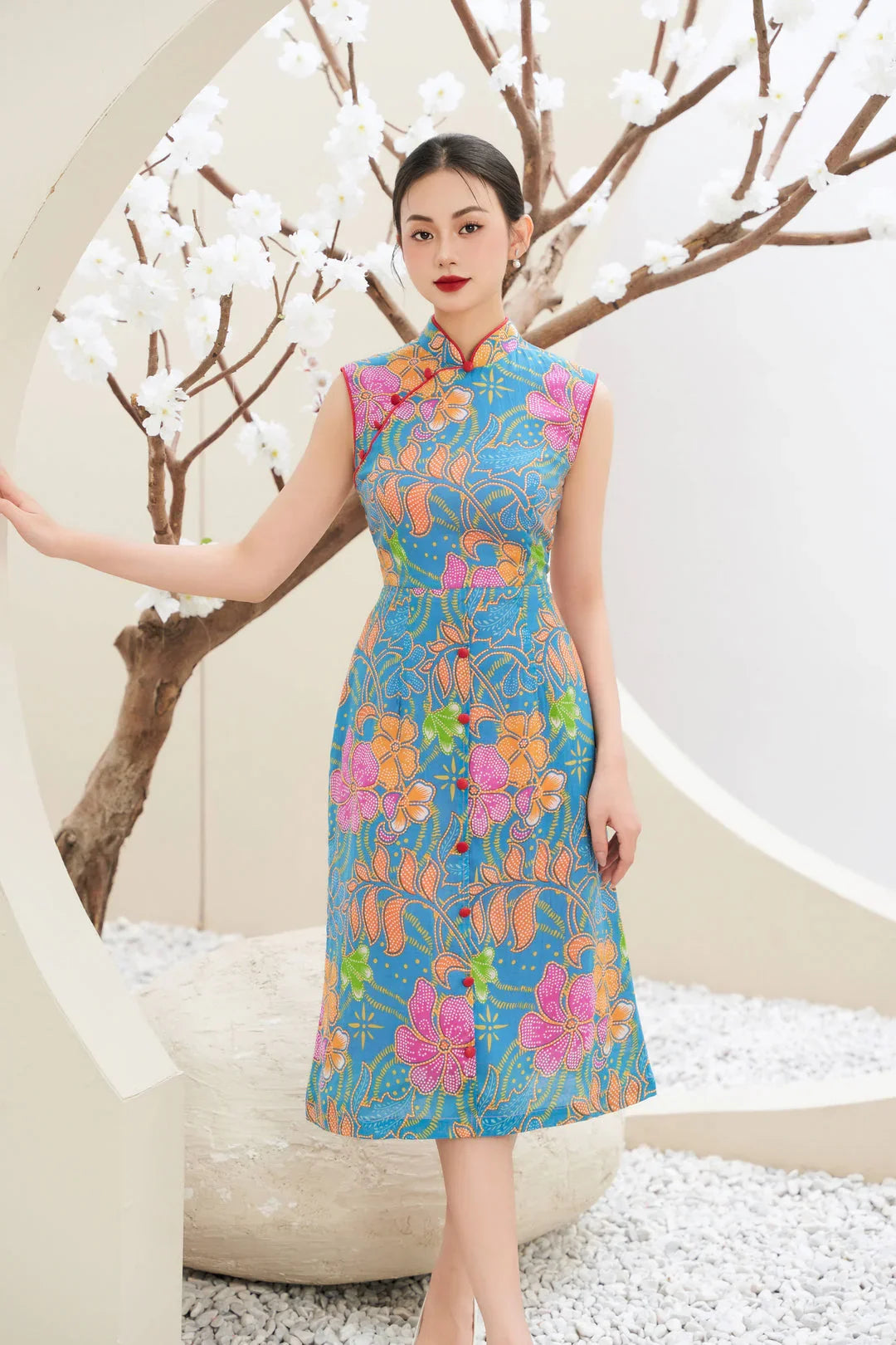 Woman in modern blue floral cheongsam dress posing by white blossom tree indoors