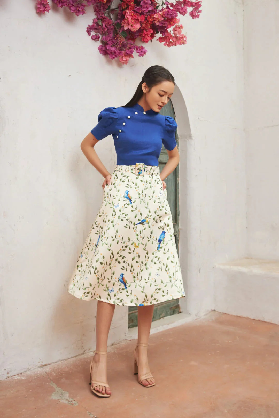 Woman in blue cheongsam top and white floral midi skirt with bird print, standing by white wall